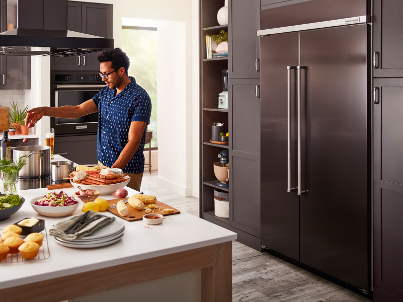 Person cooking at an island in front of a built-in, black stainless steel KitchenAid® side-by-side refrigerator Person cooking at an island in front of a built-in, black stainless steel KitchenAid® side-by-side refrigerator