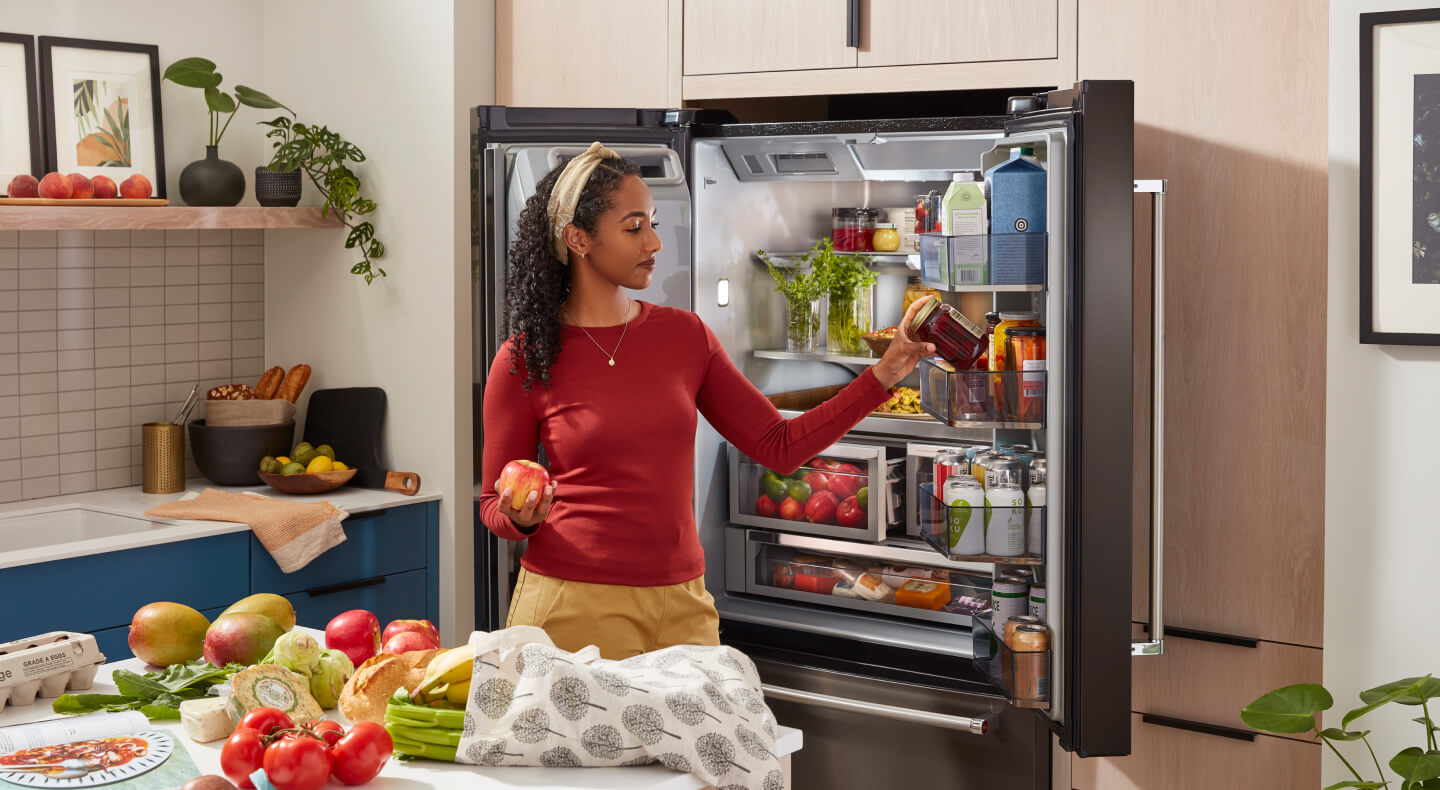 A person in a red shirt putting away groceries in a stocked French door refrigerator and a kitchen island covered in fresh produce