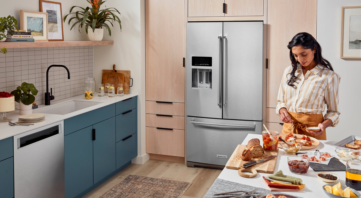 A person prepping food at an island with a bread and cheese spread in a kitchen with blue cabinets and a French door refrigerator A person prepping food at an island with a bread and cheese spread in a kitchen with blue cabinets and a French door refrigerator