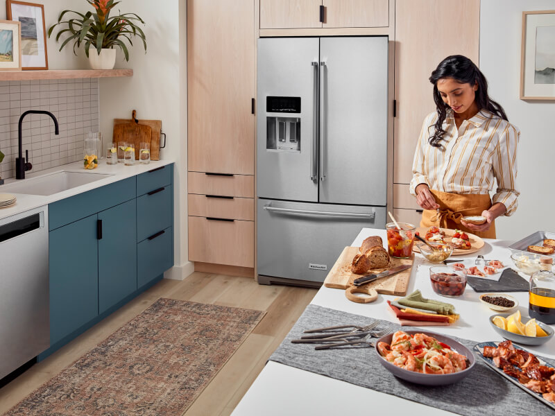 A person prepping food at an island with a bread and cheese spread in a kitchen with blue cabinets and a French door refrigerator A person prepping food at an island with a bread and cheese spread in a kitchen with blue cabinets and a French door refrigerator