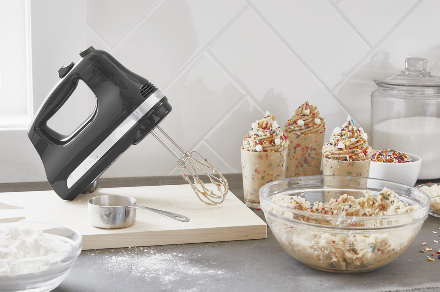 Black KitchenAid® hand mixer on wooden cutting board next to whipped cream with sprinkles in serving cups Black KitchenAid® hand mixer on wooden cutting board next to whipped cream with sprinkles in serving cups