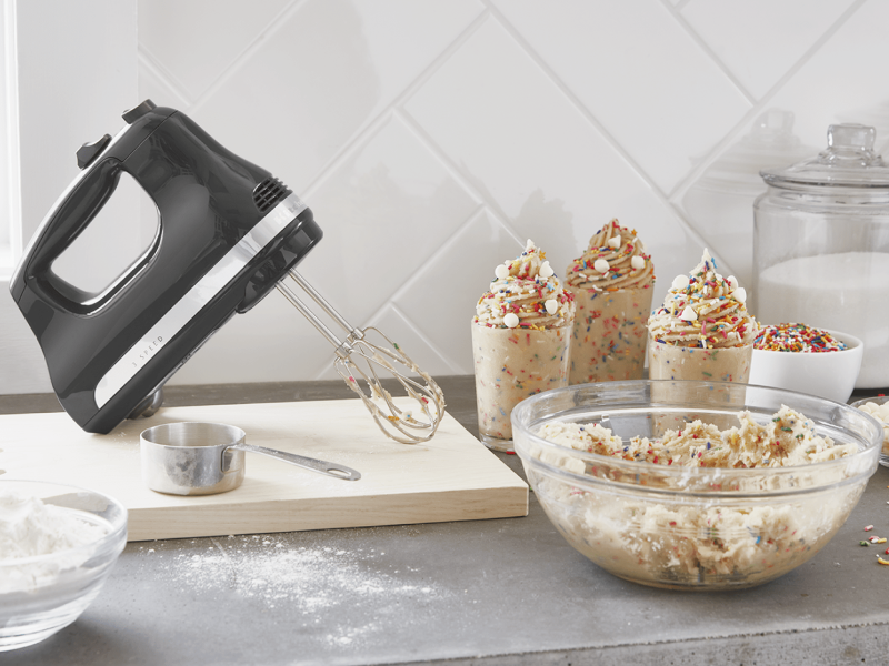 Black KitchenAid® hand mixer on wooden cutting board next to whipped cream with sprinkles in serving cups Black KitchenAid® hand mixer on wooden cutting board next to whipped cream with sprinkles in serving cups