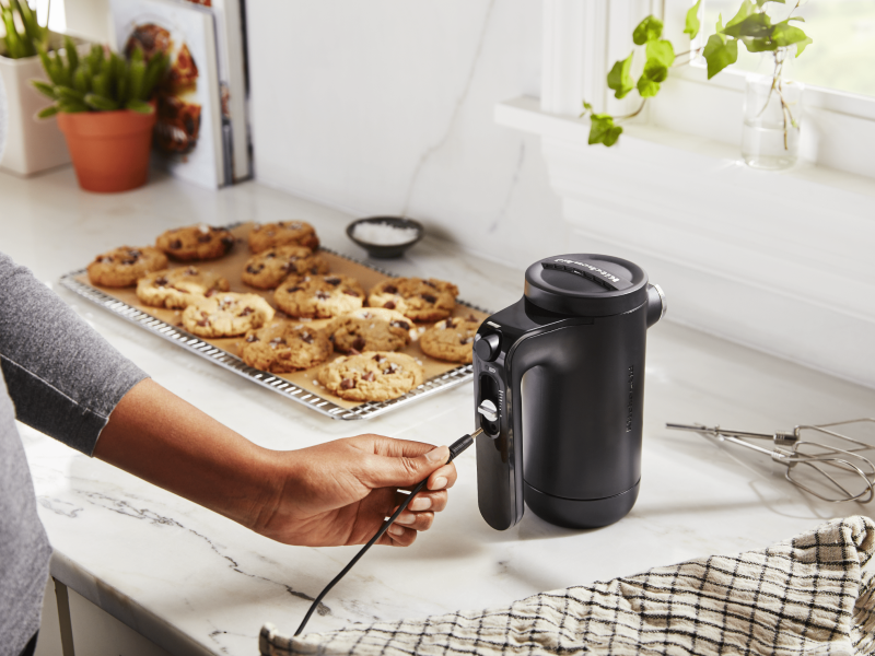 Maker plugging cord into black KitchenAid® hand mixer on countertop next to tray of chocolate chip cookies Maker plugging cord into black KitchenAid® hand mixer on countertop next to tray of chocolate chip cookies