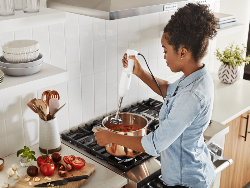 Maker using KitchenAid® immersion blender to mix ingredients in pot on top of stovetop Maker using KitchenAid® immersion blender to mix ingredients in pot on top of stovetop