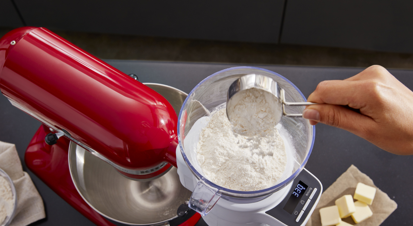 A person sifting flour into a red KitchenAid® stand mixer. A person sifting flour into a red KitchenAid® stand mixer.