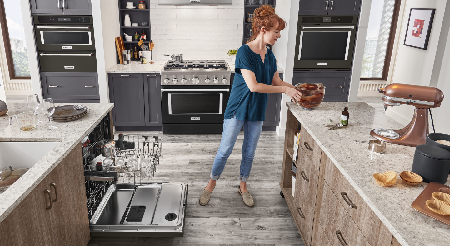 A woman placing a bowl on the counter of a modern kitchen next to a KitchenAid® dishwasher. A woman placing a bowl on the counter of a modern kitchen next to a KitchenAid® dishwasher.