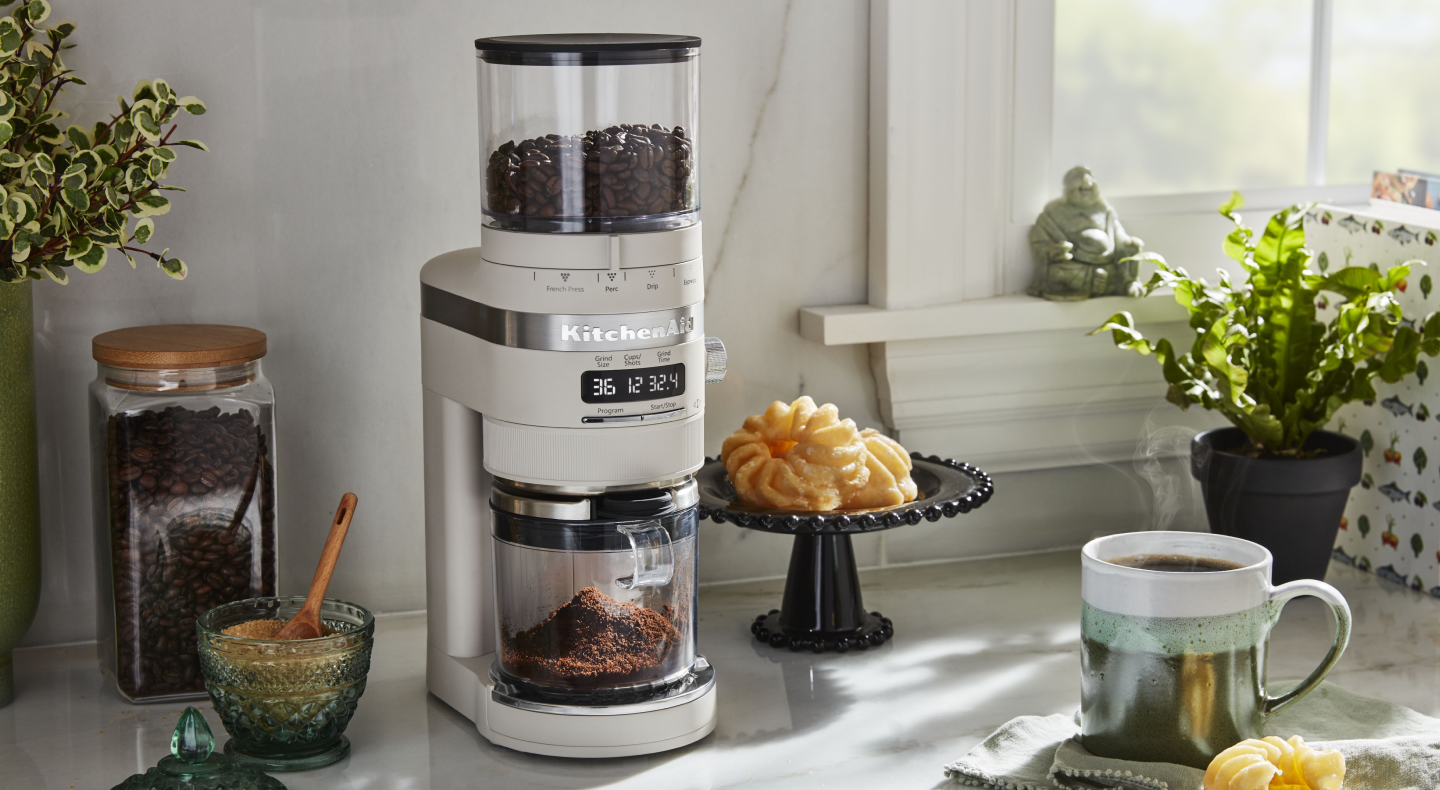 A KitchenAid® burr coffee grinder in a modern kitchen next to a pastry and cup of coffee. A KitchenAid® burr coffee grinder in a modern kitchen next to a pastry and cup of coffee.