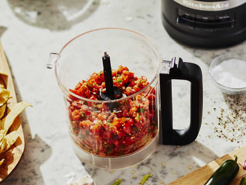 A food processor filled with salsa surrounded by the ingredients used in its creation.