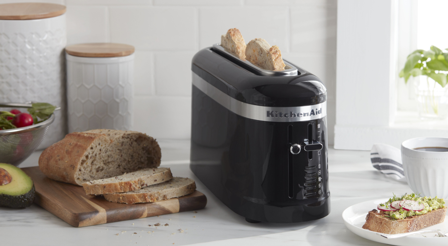 Slices of bread in a black KitchenAid® toaster next to a loaf of artisan bread and an open-faced sandwich on a modern kitchen counter. Slices of bread in a black KitchenAid® toaster next to a loaf of artisan bread and an open-faced sandwich on a modern kitchen counter.