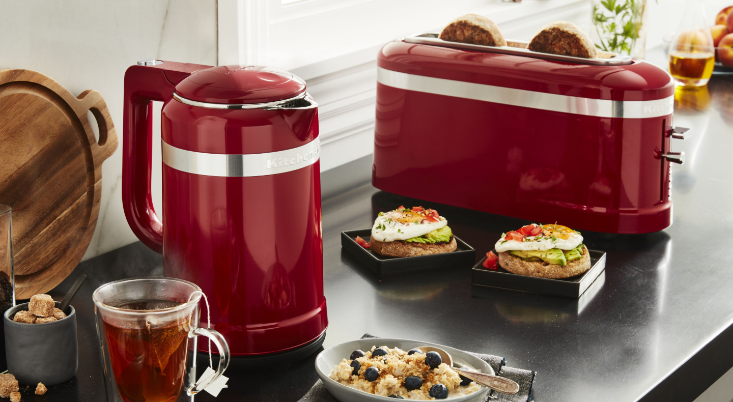 Slices of bread in a red KitchenAid® toaster next to open-faced sandwiches and cups of tea on a modern kitchen counter. Slices of bread in a red KitchenAid® toaster next to open-faced sandwiches and cups of tea on a modern kitchen counter.