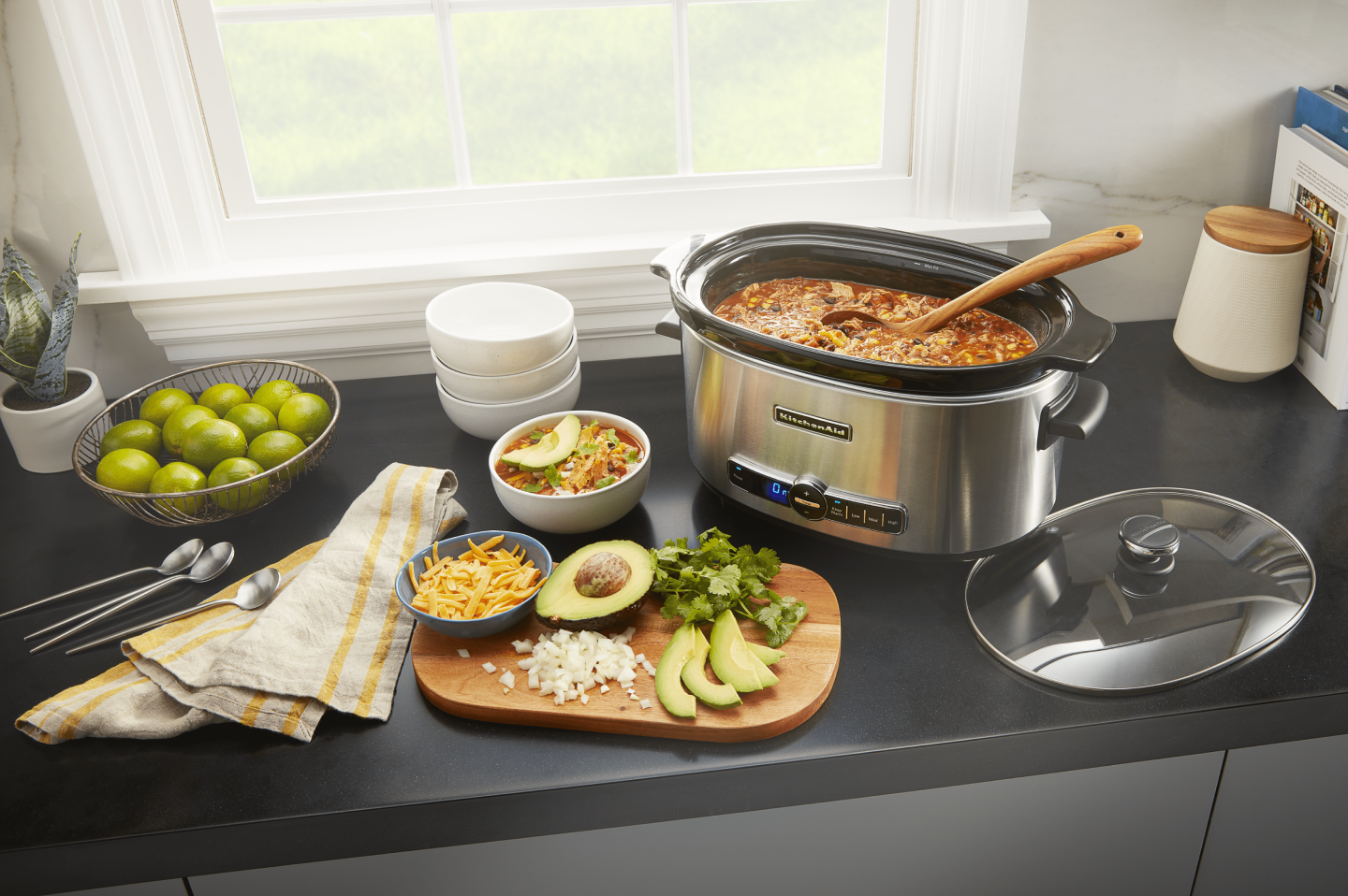 KitchenAid® slow cooker filled with chili next to a cutting board of vegetables and herbs KitchenAid® slow cooker filled with chili next to a cutting board of vegetables and herbs