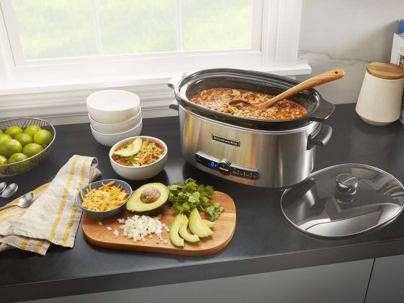 KitchenAid® slow cooker filled with chili next to a cutting board of vegetables and herbs KitchenAid® slow cooker filled with chili next to a cutting board of vegetables and herbs