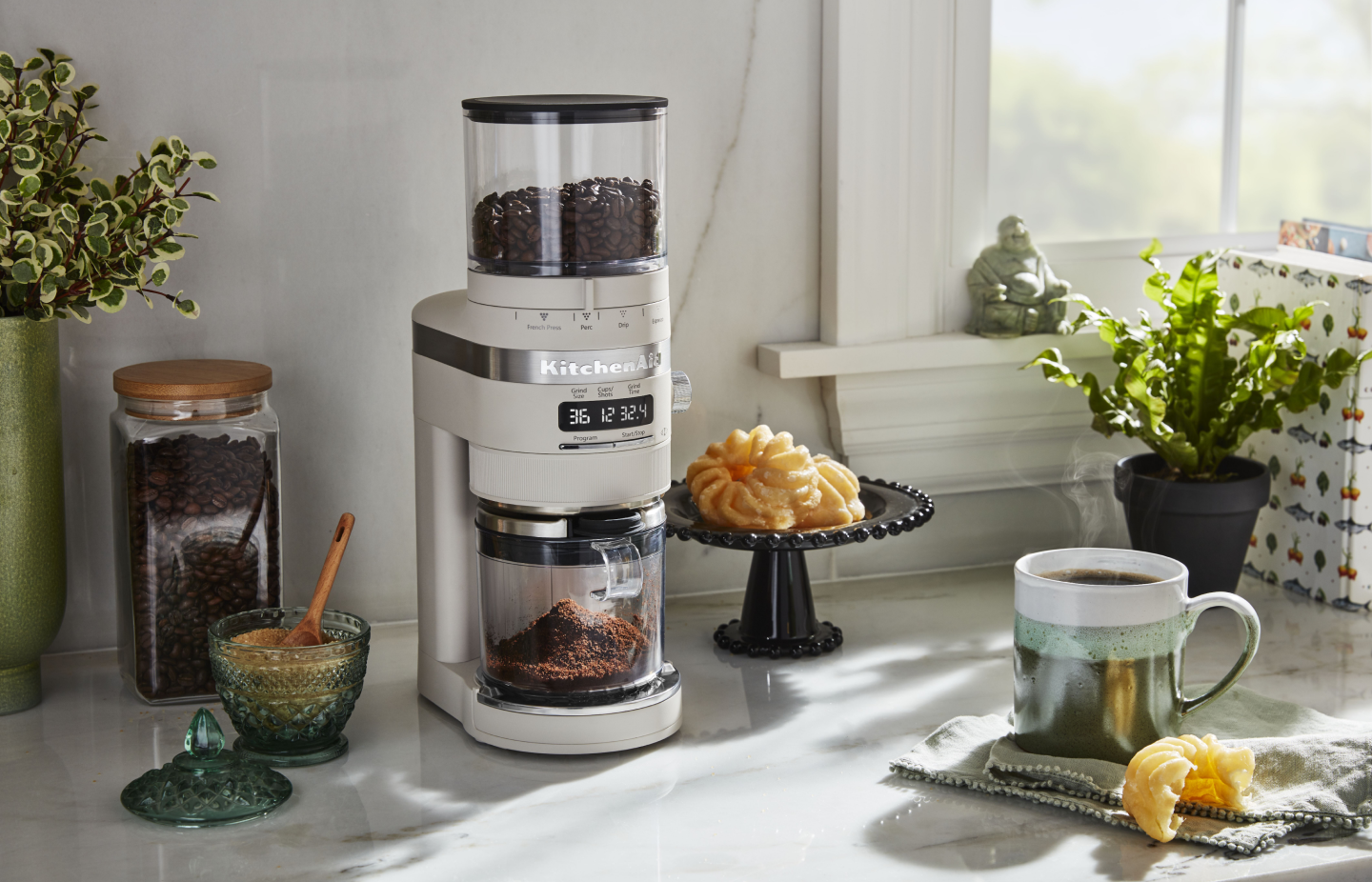 White KitchenAid® burr grinder next to a cup of coffee and pastries White KitchenAid® burr grinder next to a cup of coffee and pastries