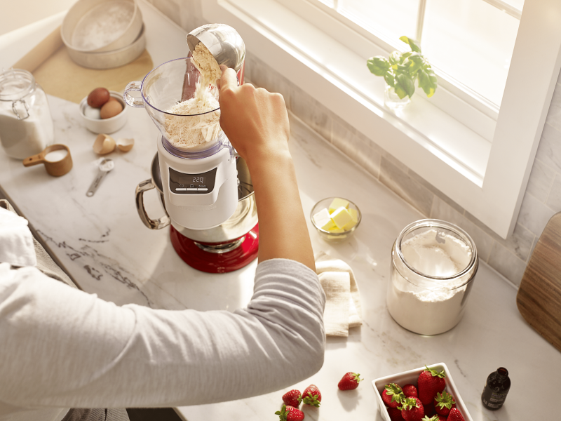 A woman adding flour to the KitchenAid®  stand mixer sifter + scale attachment.  A woman adding flour to the KitchenAid®  stand mixer sifter + scale attachment.