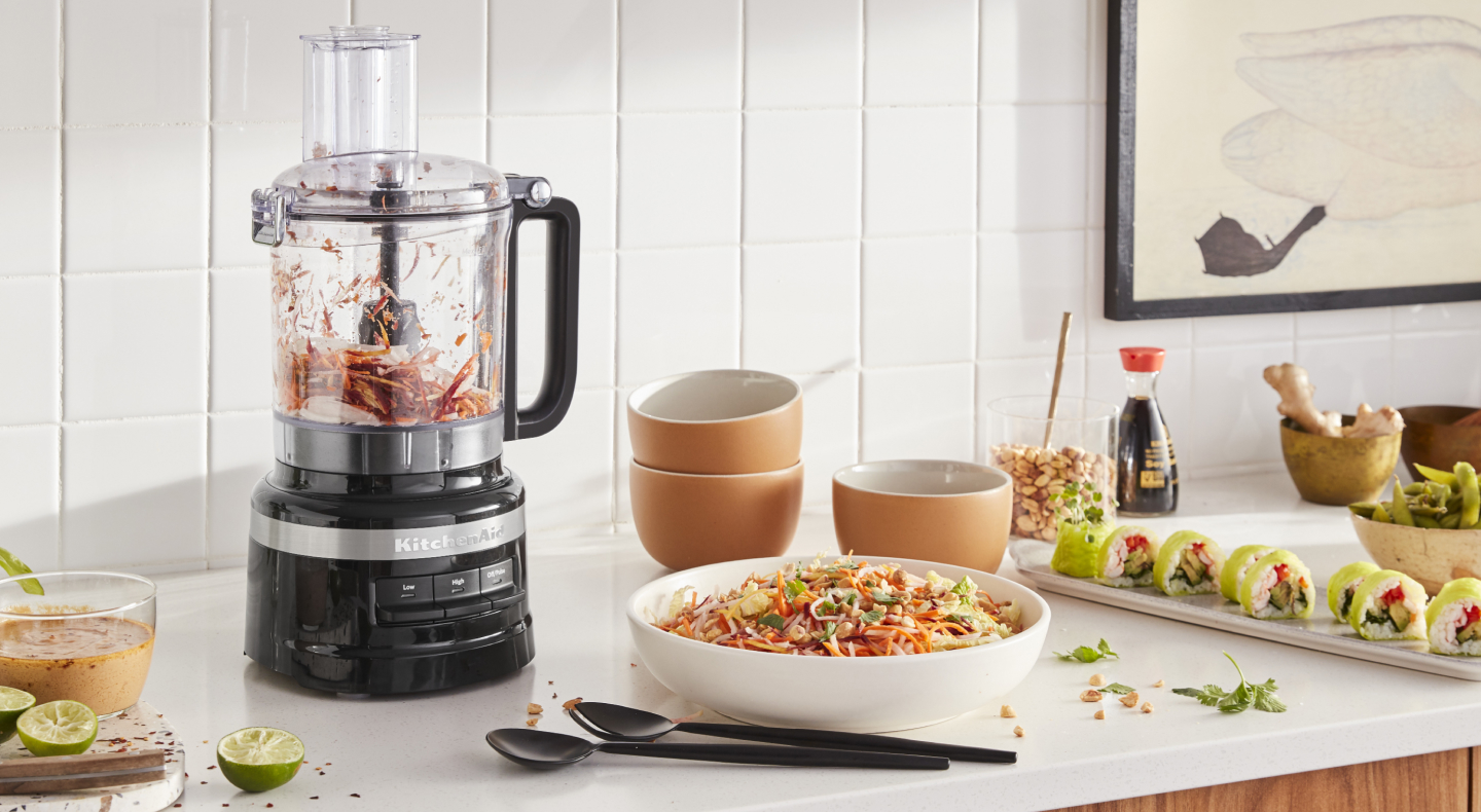 KitchenAid® food processor with chopped vegetables next to a chopped salad and freshly made salad dressing in a modern kitchen. KitchenAid® food processor with chopped vegetables next to a chopped salad and freshly made salad dressing in a modern kitchen.