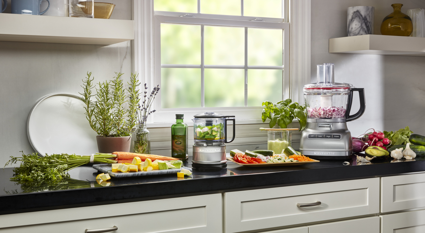 KitchenAid® food processor and food chopper with chopped vegetables next to fresh vegetables in a modern kitchen. KitchenAid® food processor and food chopper with chopped vegetables next to fresh vegetables in a modern kitchen.