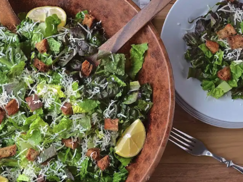 A wooden serving bowl with chopped salad next to a place setting with salad.