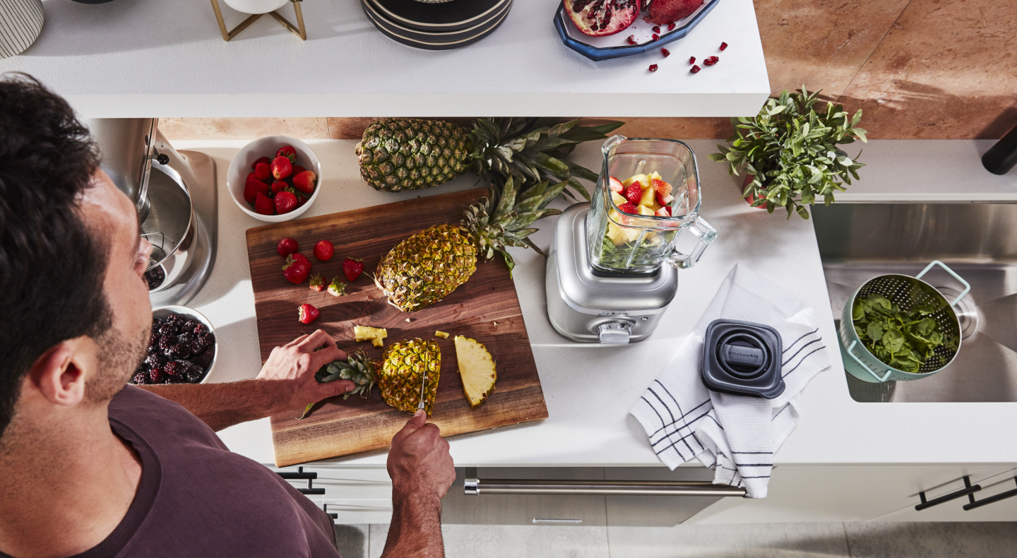 Person cutting pineapple next to KitchenAid® blender with fruit in it Person cutting pineapple next to KitchenAid® blender with fruit in it