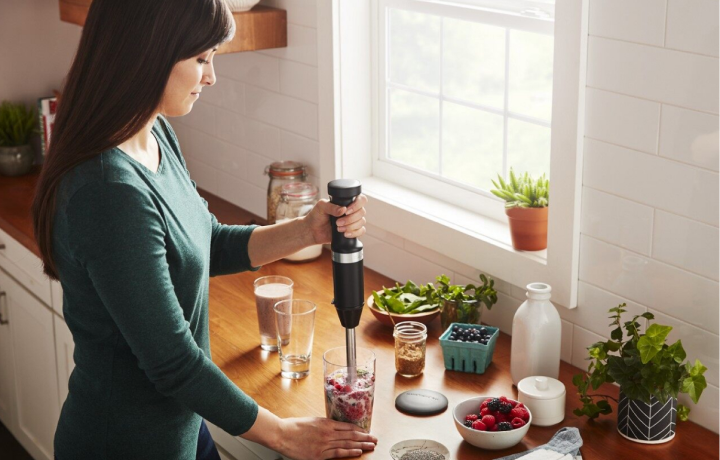 Person in green shirt blending individual smoothies with KitchenAid® immersion blender – berries, herbs and milk surround glasses