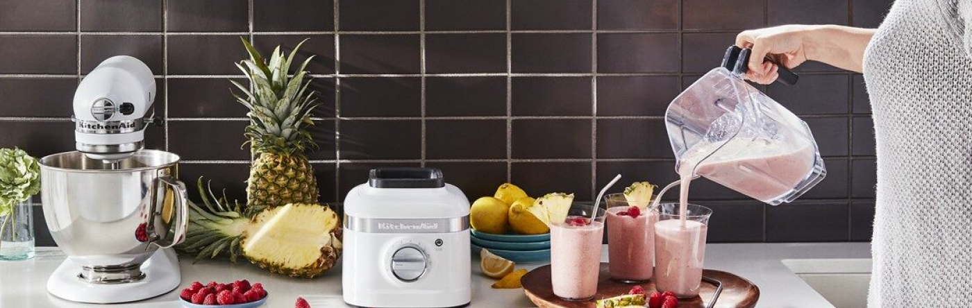 White countertop with KitchenAid® stand mixer and KitchenAid® blender surrounded by fruits - person is pouring smoothies into three glasses