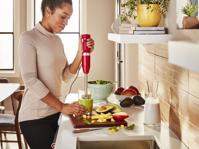 Red KitchenAid® immersion blender pureeing food in a glass next to various ingredients Red KitchenAid® immersion blender pureeing food in a glass next to various ingredients