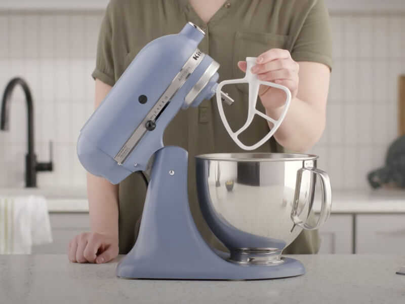 Woman holding a flat beater attachment next to a KitchenAid® stand mixer.