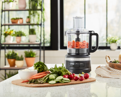 Black food processor filled with sliced carrots next to various sliced vegetables