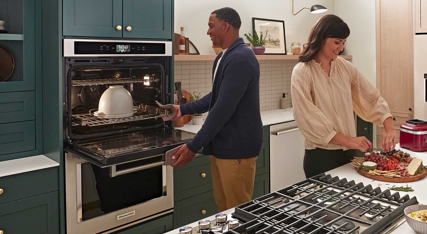 Person checking on bread in the oven while another person arranges charcuterie