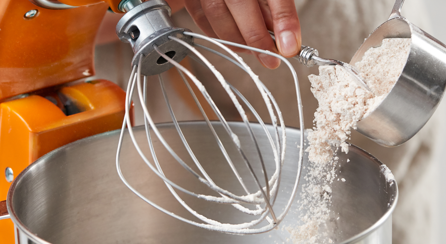 Person adding flour to stand mixer bowl beneath whip attachment Person adding flour to stand mixer bowl beneath whip attachment