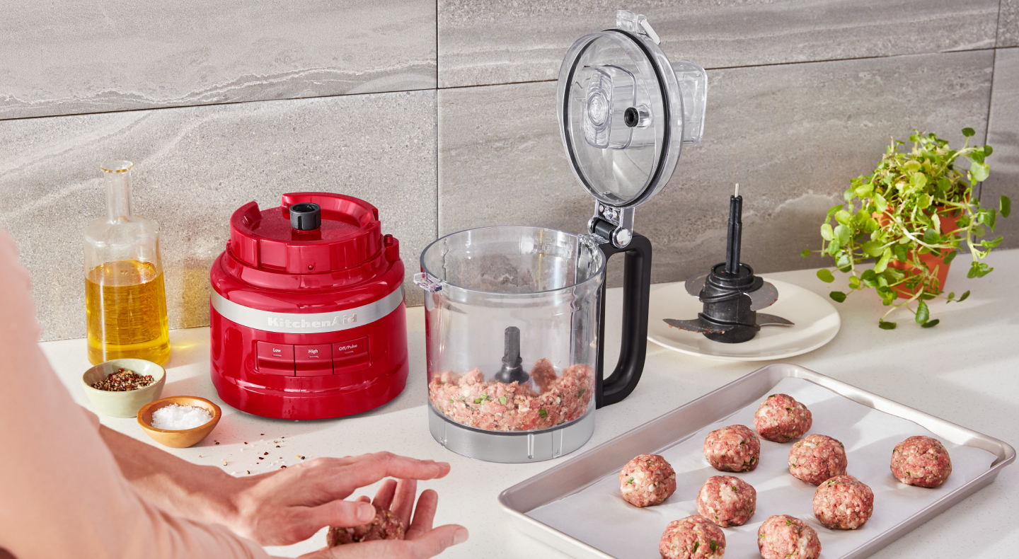 Open food processor with ground meat next to a tray of rolled ground meat and a hand rolling meatballs Open food processor with ground meat next to a tray of rolled ground meat and a hand rolling meatballs