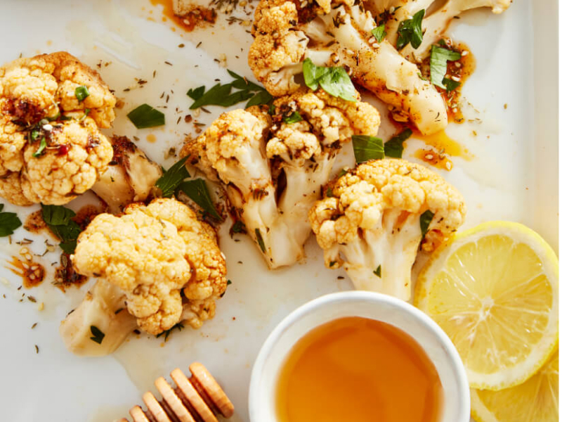 Honey roasted cauliflower with small pot of honey and lemon slice in foreground.