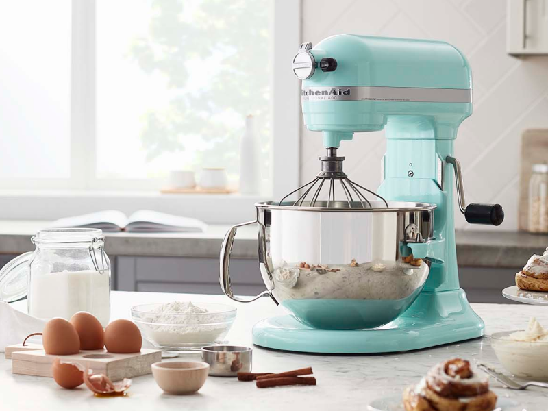KitchenAid® stand mixer with a wire whip accessory next to cracked egg shells and fresh baked cinnamon rolls.