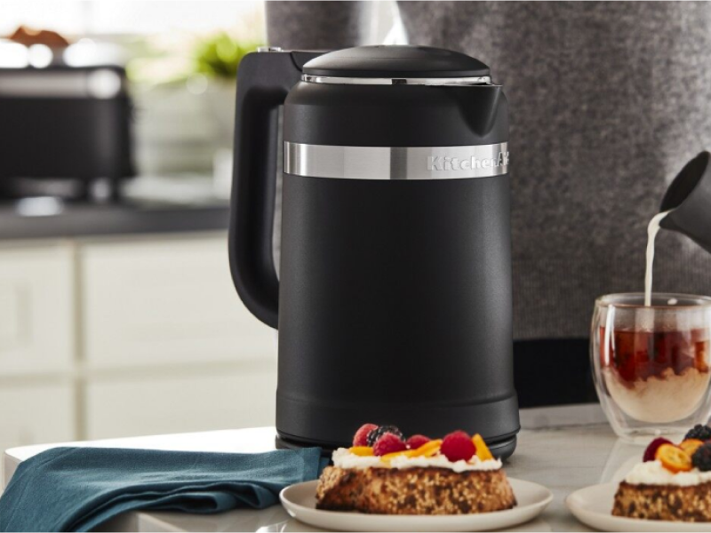 Hand pouring milk into cups of tea sitting next to electric tea kettle and plates of pastries.
