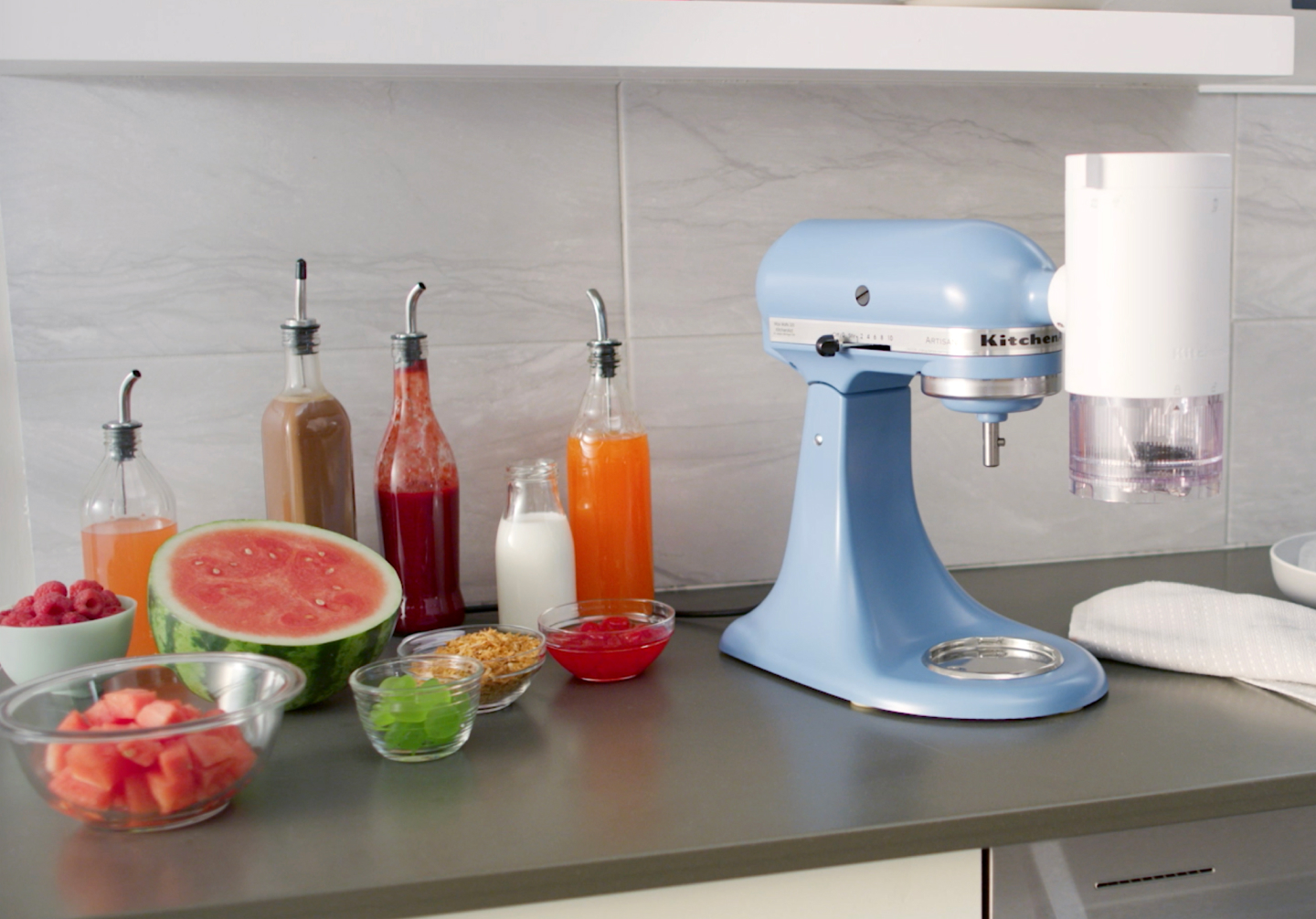 Blue stand mixer with KitchenAid® Shave Ice Attachment on counter with shaved ice syrups, watermelon and ingredients Blue stand mixer with KitchenAid® Shave Ice Attachment on counter with shaved ice syrups, watermelon and ingredients