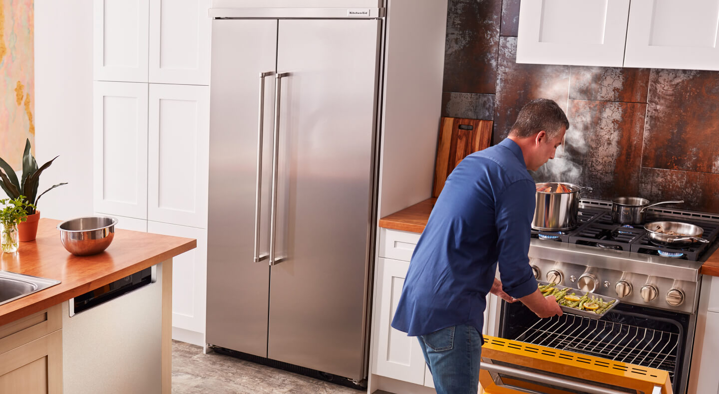 Person loading food into a gas KitchenAid® oven next to a stainless steel side-by-side refrigerator Person loading food into a gas KitchenAid® oven next to a stainless steel side-by-side refrigerator