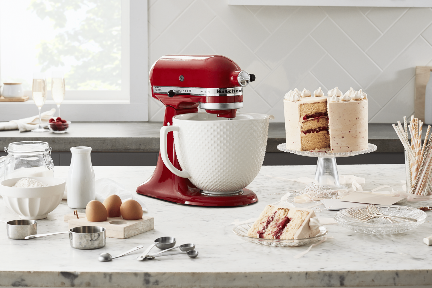 Red KitchenAid® Stand Mixer on a counter next to a three layer white frosted cake, milk, eggs, measuring cups and flour  Red KitchenAid® Stand Mixer on a counter next to a three layer white frosted cake, milk, eggs, measuring cups and flour