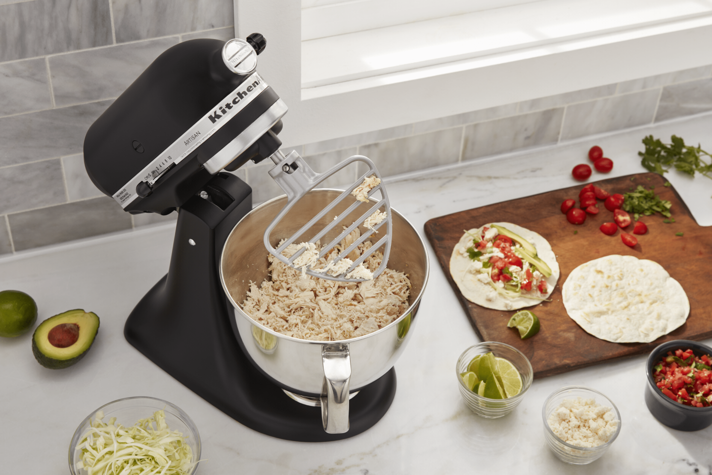 Black KitchenAid® Stand Mixer shredding chicken using the paddle attachment while surrounded by tortillas, salsa and taco fixings on a counter  Black KitchenAid® Stand Mixer shredding chicken using the paddle attachment while surrounded by tortillas, salsa and taco fixings on a counter