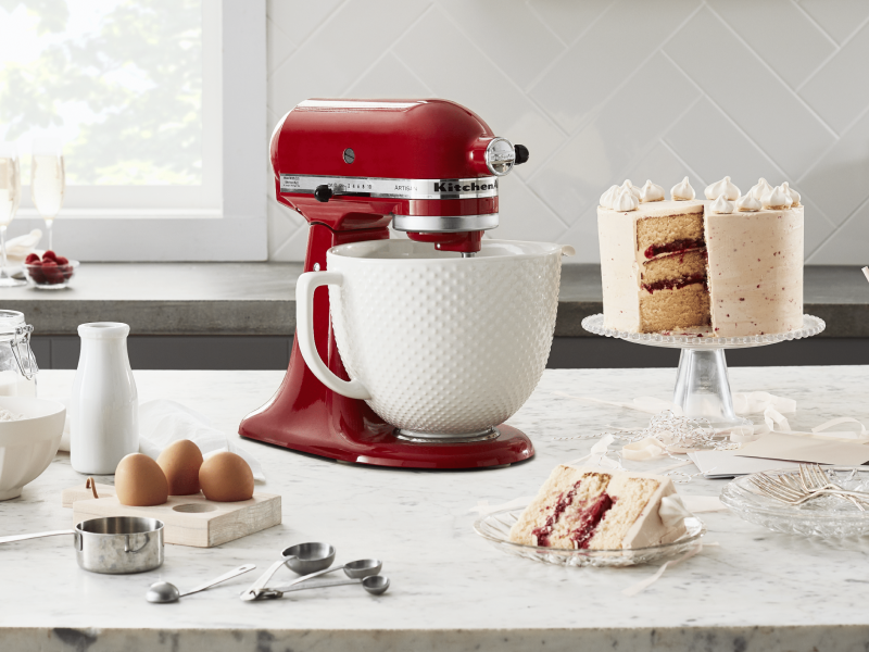 Red KitchenAid® Stand Mixer on a counter next to a three layer white frosted cake, milk, eggs, measuring cups and flour  Red KitchenAid® Stand Mixer on a counter next to a three layer white frosted cake, milk, eggs, measuring cups and flour
