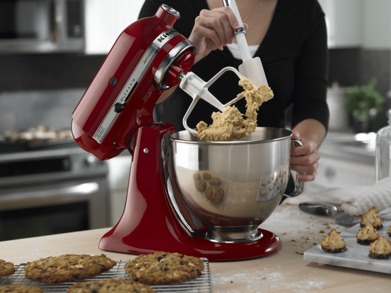 Tilt-head red KitchenAid® Stand Mixer with paddle attachment being scraped down while mixing up cookie dough with baked cookies in the foreground