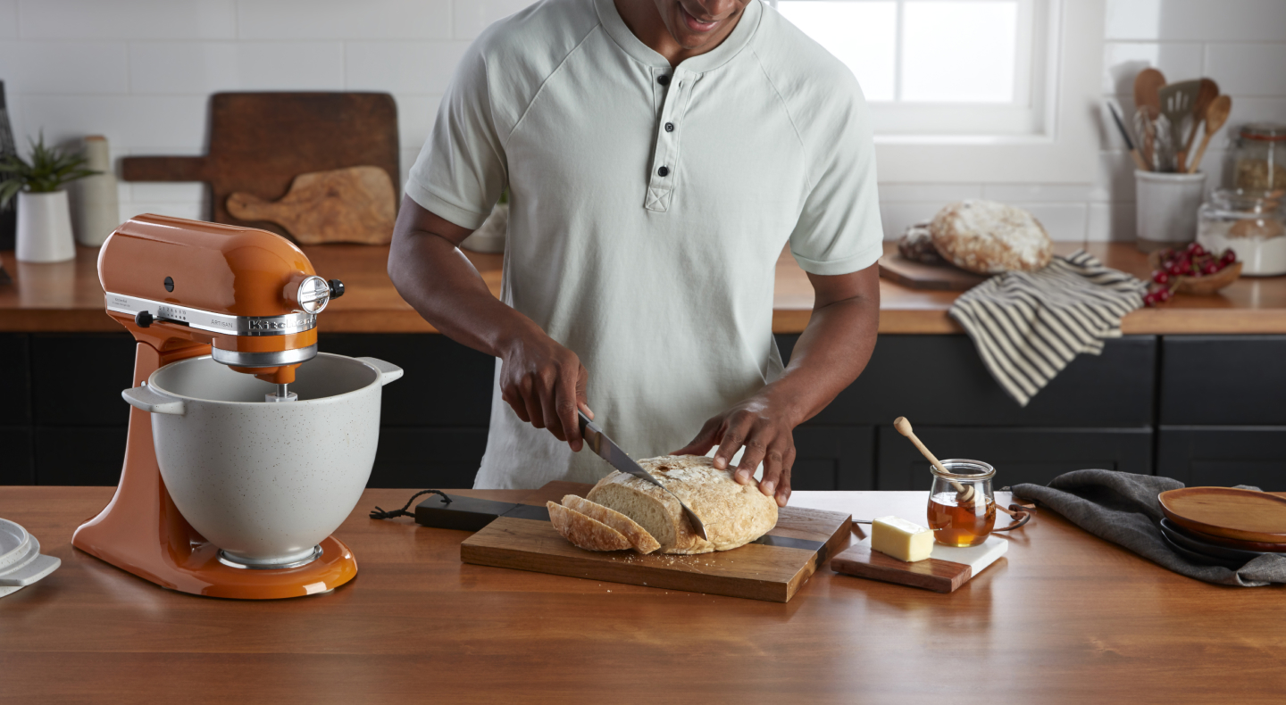 Person slicing loaf of bread on a cutting board next to an orange stand mixer