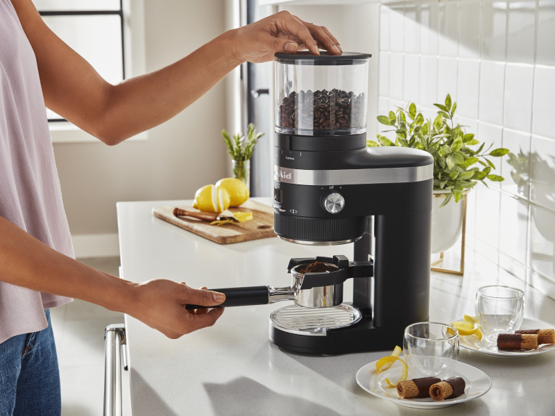 A woman using a black KitchenAid® Burr Coffee Grinder to process coffee beans A woman using a black KitchenAid® Burr Coffee Grinder to process coffee beans
