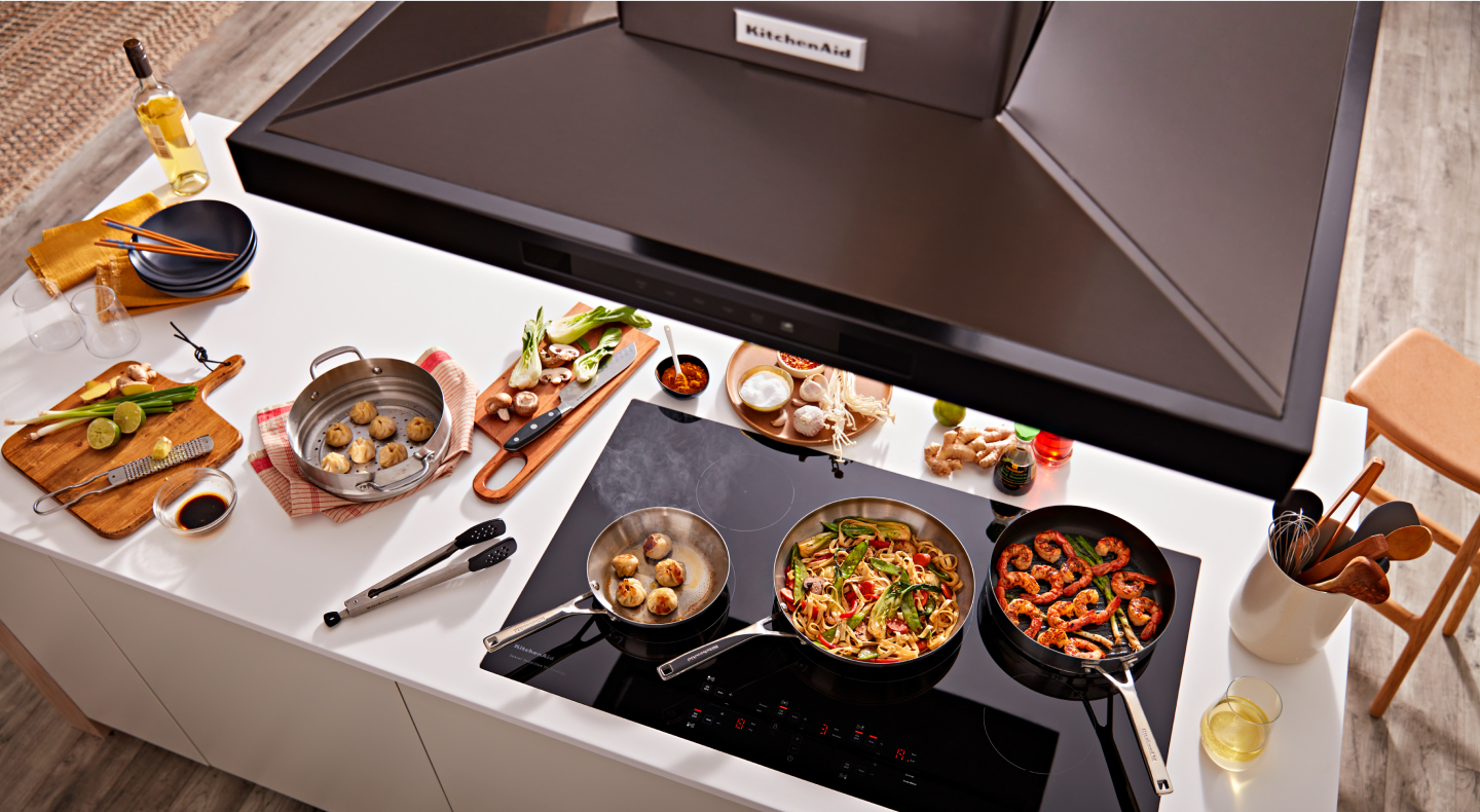 Overhead view of KitchenAid® range hood and pans with food on black cooktop Overhead view of KitchenAid® range hood and pans with food on black cooktop
