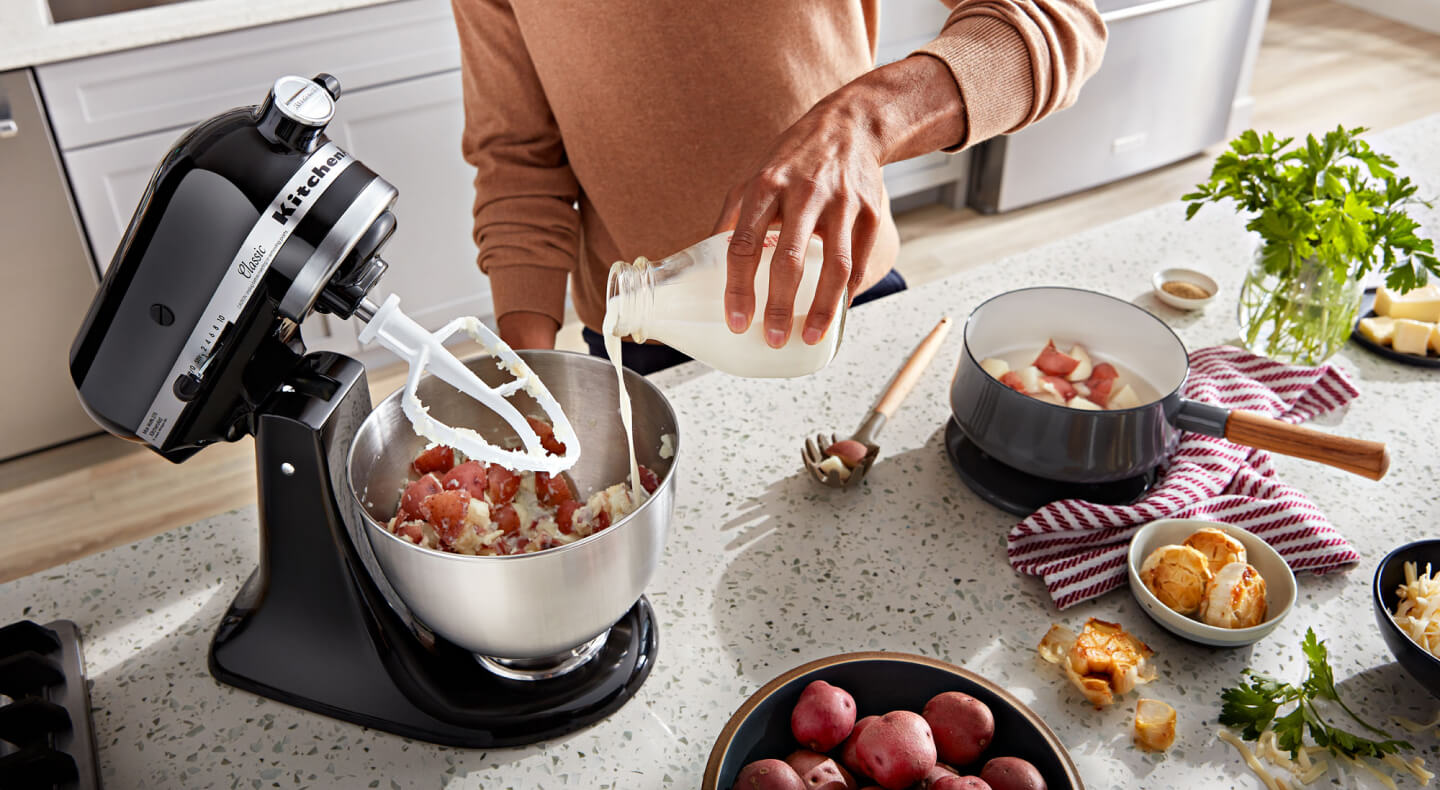 A person pouring milk into a KitchenAid® stand mixer bowl with ingredients and a flat beater accessory on the counter of a modern kitchen. A person pouring milk into a KitchenAid® stand mixer bowl with ingredients and a flat beater accessory on the counter of a modern kitchen.