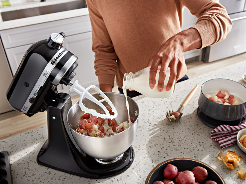 A person pouring milk into a KitchenAid® stand mixer bowl with ingredients and a flat beater accessory on the counter of a modern kitchen. A person pouring milk into a KitchenAid® stand mixer bowl with ingredients and a flat beater accessory on the counter of a modern kitchen.