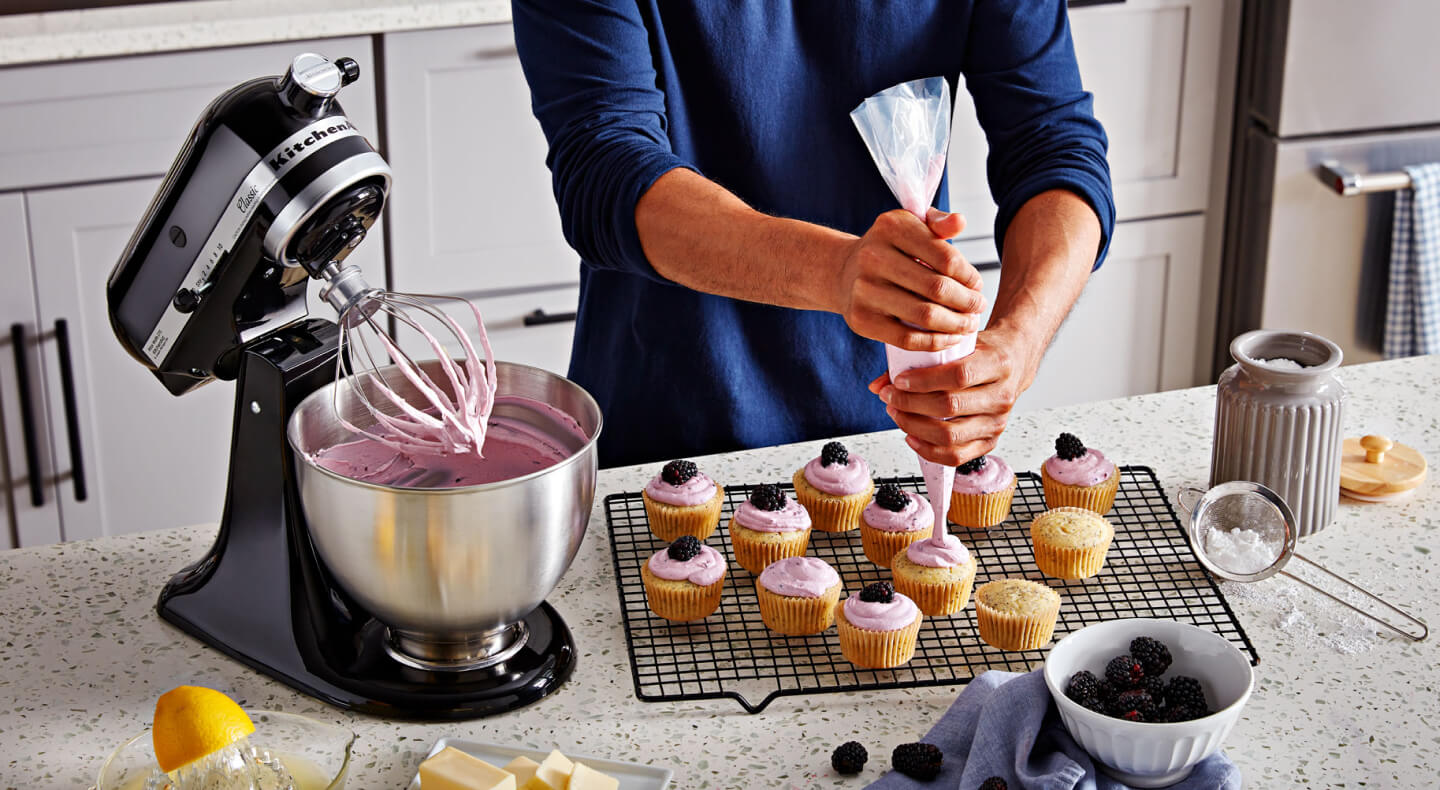 A person frosting cupcakes on the counter of a modern kitchen next to a KitchenAid® stand mixer with a wire whisk/whip accessory. A person frosting cupcakes on the counter of a modern kitchen next to a KitchenAid® stand mixer with a wire whisk/whip accessory.