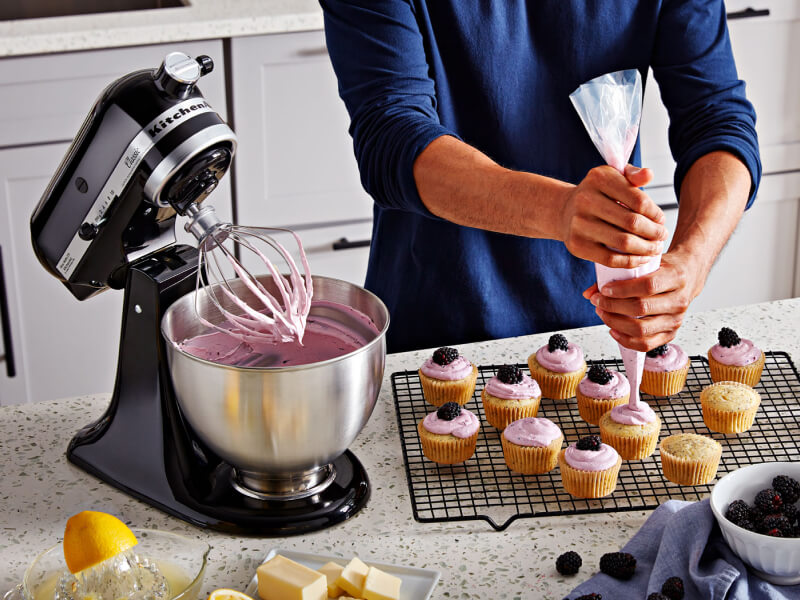 A person frosting cupcakes on the counter of a modern kitchen next to a KitchenAid® stand mixer with a wire whisk/whip accessory. A person frosting cupcakes on the counter of a modern kitchen next to a KitchenAid® stand mixer with a wire whisk/whip accessory.