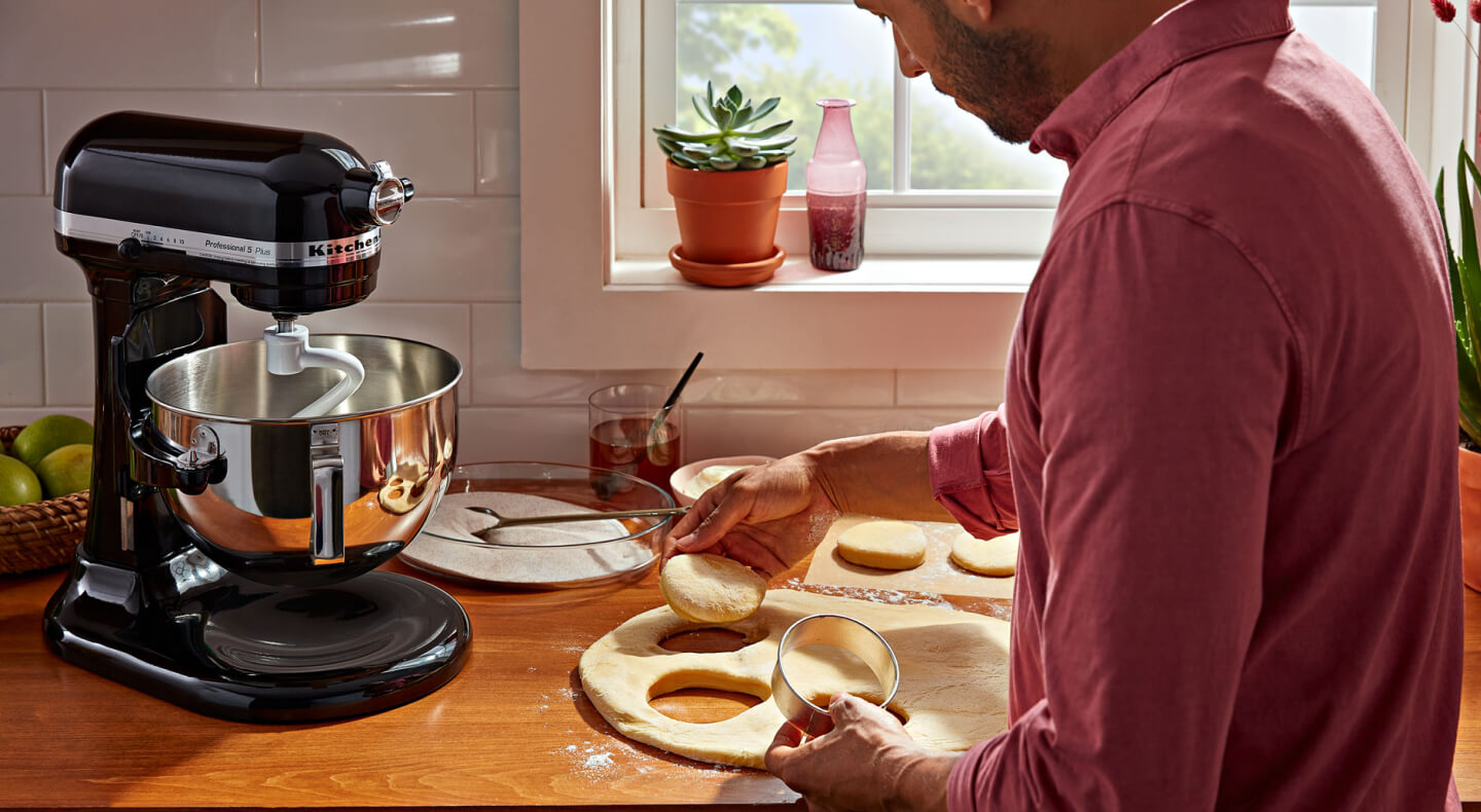A person cutting biscuit dough on the counter of a modern kitchen next to a KitchenAid® stand mixer with a dough hook accessory. A person cutting biscuit dough on the counter of a modern kitchen next to a KitchenAid® stand mixer with a dough hook accessory.