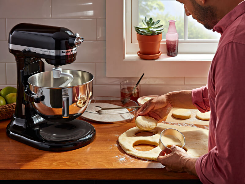 A person cutting biscuit dough on the counter of a modern kitchen next to a KitchenAid® stand mixer with a dough hook accessory. A person cutting biscuit dough on the counter of a modern kitchen next to a KitchenAid® stand mixer with a dough hook accessory.