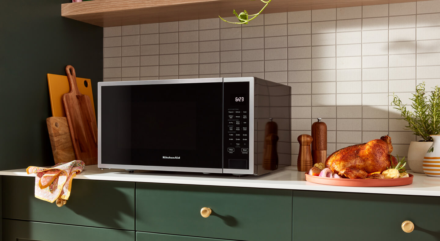 A KitchenAid® countertop microwave on a white counter with green cabinets and a roasted chicken next to it
