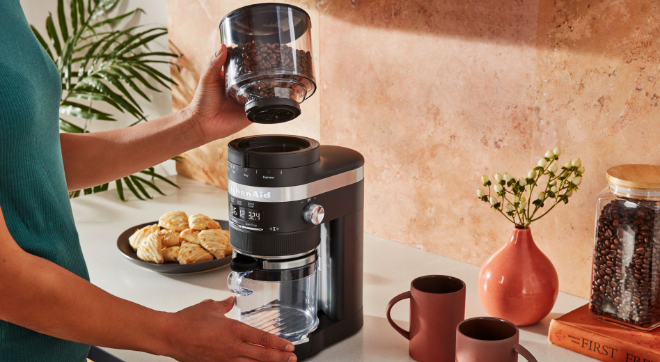 A woman using a black KitchenAid® coffee grinder to grind coffee beans A woman using a black KitchenAid® coffee grinder to grind coffee beans